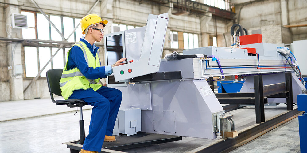 Concentrated operator wearing reflective jacket and hardhat focused on work while sitting in front of CNC machine, interior of spacious production department on background