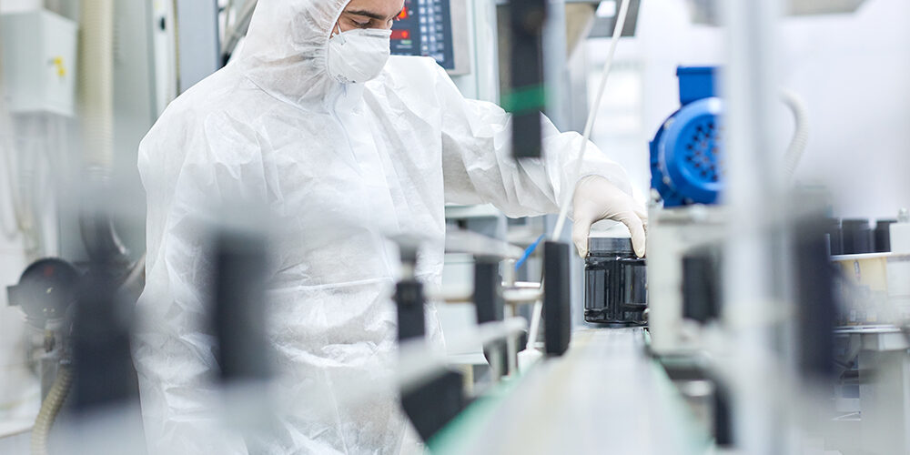 Young man wearing coverall and safety mask working on production line of modern pharmaceutical factory, portrait shot